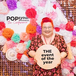 Bec holding a sign "THE event of the year" in front of a backdrop of colourful flowers and a sign that says "Pop Your Business The Conference"
